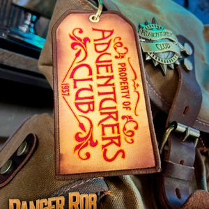 May include: A brown leather patch with the text "ADVENTURERS CLUB 1957" in red and gold lettering. The patch is attached to a brown leather bag with a metal buckle. The bag has a metal tag with the text "ADVENTUREX CLUB" in gold lettering.