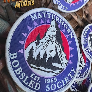 May include: A blue and white circular patch with the text "Matterhorn Bobsled Society Est. 1959" and a white mountain illustration with a red and white sky.