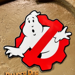 May include: A red and white sticker with a ghost figure inside a gear shape with a diagonal red line through it. The sticker has a black outline and is on a brown background. The text "RANGER ROB ARTifACTS" is below the sticker.