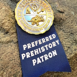 May include: A gold-colored metal badge from "The Dino Institute" with a blue ribbon that reads "Preferred Prehistory Patron." The badge features a triceratops and other prehistoric symbols. The badge is set against a textured rock background.