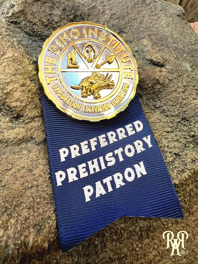 May include: A gold-colored metal badge from "The Dino Institute" with a blue ribbon that reads "Preferred Prehistory Patron." The badge features a triceratops and other prehistoric symbols. The badge is set against a textured rock background.