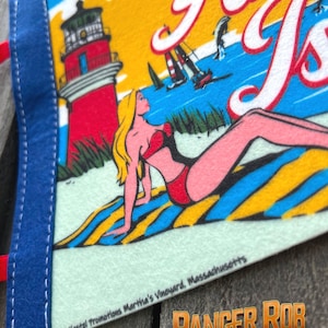 May include: A vintage pennant featuring a beach scene with the words "Anna's Isle". The pennant has a blue border and a red lighthouse, a woman in a red swimsuit, and sailboats on the water. The text "1975 Kastel Promotions Martha's Vineyard, Massachusetts" is also visible.