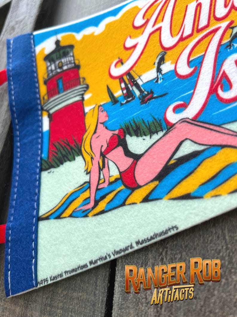 May include: A vintage pennant featuring a beach scene with the words "Anna's Isle". The pennant has a blue border and a red lighthouse, a woman in a red swimsuit, and sailboats on the water. The text "1975 Kastel Promotions Martha's Vineyard, Massachusetts" is also visible.
