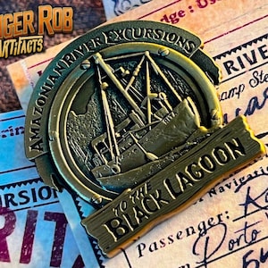May include: A gold-colored metal medallion with the text "Amazonian River Excursions" and "To the Black Lagoon". The medallion features a small boat with the word "Rita" on the side. The medallion is resting on a piece of paper with the text "Passenger: Robert Porto Velho 54" written on it.