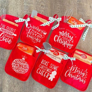 Può includere: Set di sei guanti da forno rossi con testo e disegni bianchi. I guanti hanno tasche per piccoli regali, come miscele per la cottura, e sono legati con un nastro verde e bianco. Il testo sui guanti include "The Secret Ingredient is Always Love", "Whiskey is my Love Language", "Have Yourself a Merry Little Christmas", "Happy Holidays", "Bake Some Christmas Cheer" e "Merry Christmas".