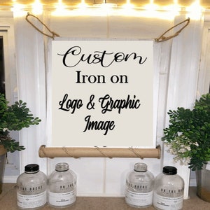 May include: A rectangular sign with the words "Custom Iron on Logo & Graphic Image" in black script. The sign is cream-colored and suspended by rope. Decorative bottles and greenery are in the foreground.