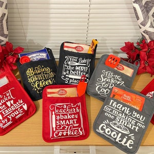 Op de afbeelding: Set van 7 rode, zwarte en grijze ovenwantjes met verschillende spreuken met een kerst- en leerkrachtthema. De wantjes hebben zakken om kleine items zoals bakmixen of keukengerei in te bewaren.