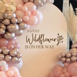 以下が含まれることがあります： 白い背景に、茶色の文字で「A little Wildflower is on her way」と書かれています。背景はピンクとゴールドの風船で飾られています。