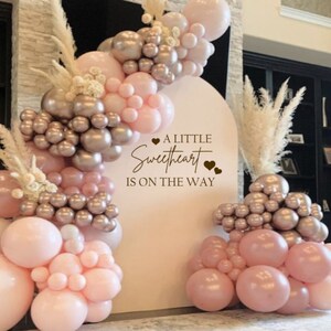 May include: A white backdrop with a pink and gold balloon arch and the text "A little sweetheart is on the way".