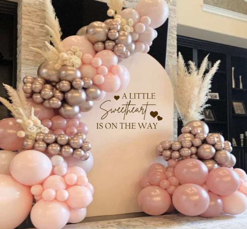May include: A white backdrop with a pink and gold balloon arch and the text "A little sweetheart is on the way".