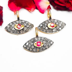 May include: Three eye-shaped pendants with gold bails, set with small, sparkling stones and a central red gemstone. The pendants are arranged on a white surface, with red rose petals in the background.