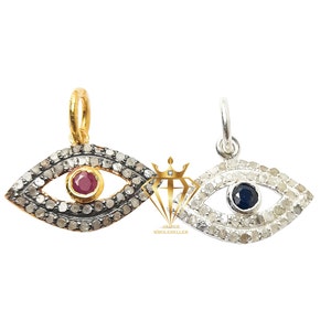Puede incluir: Dos colgantes con forma de ojo. Uno es dorado con un centro de rubí y el otro es plateado con un centro azul oscuro. Ambos están rodeados de pequeñas piedras transparentes. Los colgantes están sobre un fondo blanco.