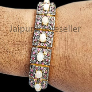 Puede incluir: Un brazalete de oro con una fila de ópalos ovalados engastados en marcos cuadrados. El brazalete está adornado con pequeñas piedras preciosas rosas y transparentes.