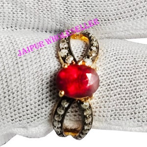 May include: A gold-toned ring with a central oval-shaped red gemstone. The ring features a unique infinity-style design with small, sparkling accents. The ring is displayed on a white surface.