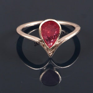 May include: A rose gold ring with a pear-shaped, vibrant red gemstone. The ring's band forms a V-shape below the gemstone, creating a unique and elegant design. The ring is set against a dark background, highlighting its reflective qualities.