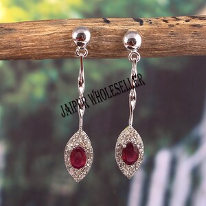 Peut inclure: Une paire de boucles d'oreilles pendantes en argent avec une pierre de rubis ovale rouge entourée d'un halo de petites pierres blanches. Les boucles d'oreilles ont un design torsadé et une boule d'argent en haut.
