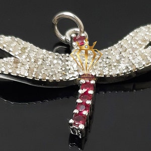 Puede incluir: Colgante de plata con forma de libélula. Las alas están incrustadas con piedras similares a diamantes, y el cuerpo presenta una línea de piedras preciosas rojas. El colgante tiene un lazo para una cadena, sobre fondo negro.