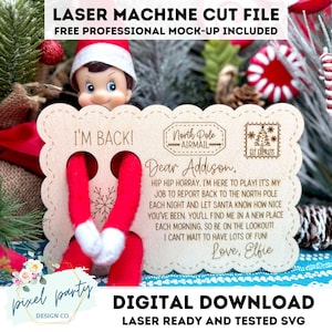 Puede incluir: Decoración navideña de madera cortada con láser con el texto "I'M BACK!" y un mensaje a "Addison" de "Elfie". La decoración presenta un copo de nieve y un sello de North Pole Airmail. Una muñeca elfo es visible.
