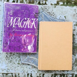 May include: Two books on a weathered surface. One book has a purple cover with the word "MAGICK" in white, along with other text and graphics. The other book has a plain tan cover. The books are resting on a decorative, grey, star-shaped object.