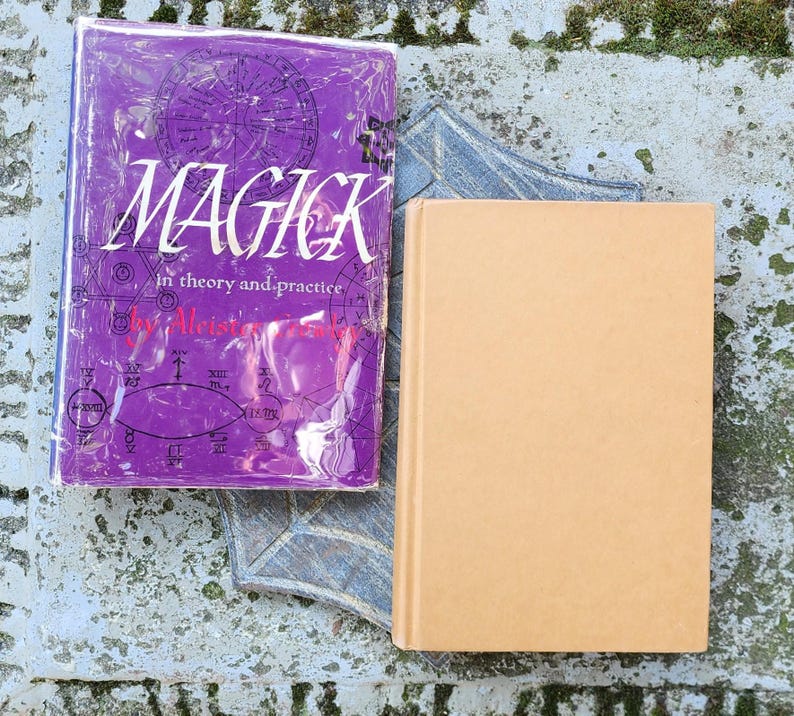 May include: Two books on a weathered surface. One book has a purple cover with the word "MAGICK" in white, along with other text and graphics. The other book has a plain tan cover. The books are resting on a decorative, grey, star-shaped object.