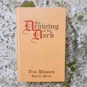 May include: A hardcover book titled "The Drawing of the Dark" by Tim Powers, published by Hypatia Press. The cover is a light brown with the title in ornate red lettering. The book is set against a textured, light-colored background.