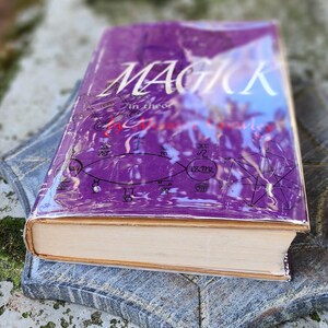 May include: A purple hardcover book titled "MAGICK" with white text, displayed on a grey stone surface. The book's cover is adorned with symbols and is encased in clear plastic.