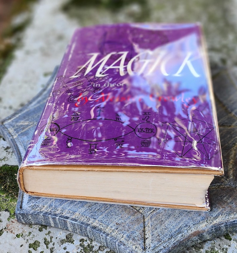May include: A purple hardcover book titled "MAGICK" with white text, displayed on a grey stone surface. The book's cover is adorned with symbols and is encased in clear plastic.