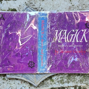 May include: A book titled "MAGICK in theory and practice" by Aleister Crowley. The book has a purple cover with white text and illustrations, and is wrapped in clear plastic. The spine is blue with the author's name. The book is open, revealing the cover and the back cover.