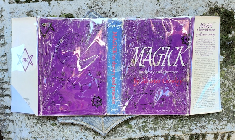 May include: A book titled "MAGICK in theory and practice" by Aleister Crowley. The book has a purple cover with white text and illustrations, and is wrapped in clear plastic. The spine is blue with the author's name. The book is open, revealing the cover and the back cover.
