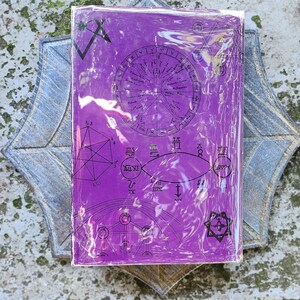 May include: A purple book with astrological symbols and a circular chart design, wrapped in clear plastic. The book rests on a grey, star-shaped decorative element, creating a mystical aesthetic. The design includes geometric shapes.