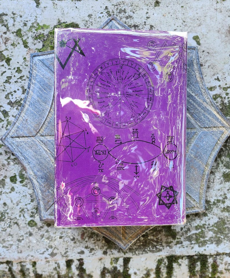 May include: A purple book with astrological symbols and a circular chart design, wrapped in clear plastic. The book rests on a grey, star-shaped decorative element, creating a mystical aesthetic. The design includes geometric shapes.