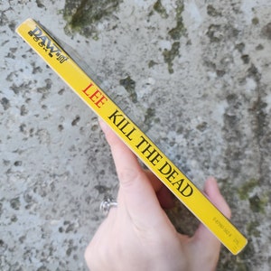 May include: A yellow paperback book titled "KILL THE DEAD" by Lee. The book's spine features the author's name and the publisher's logo in black and red. The book is held against a textured, grey background.