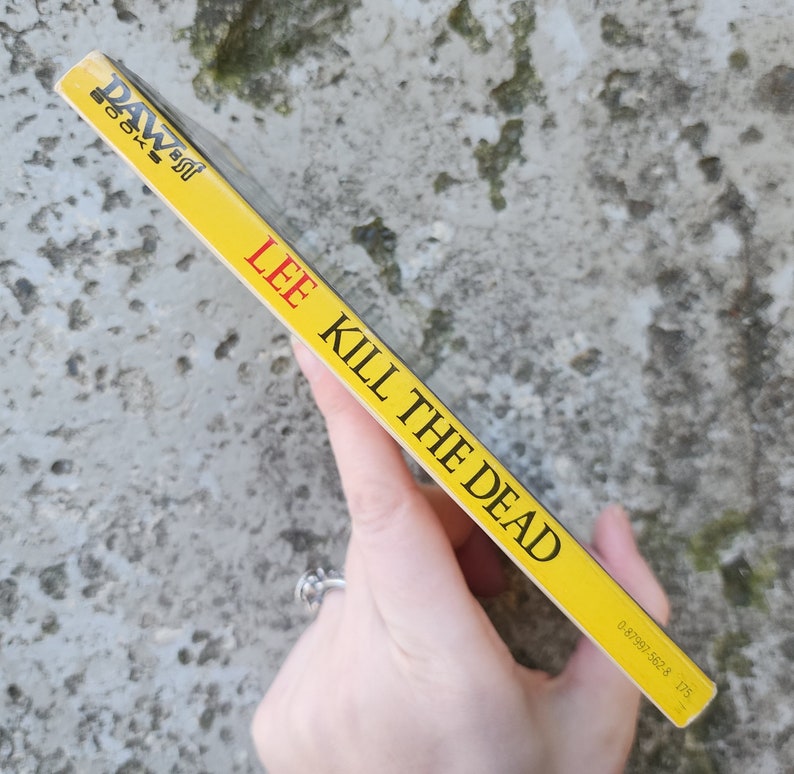 May include: A yellow paperback book titled "KILL THE DEAD" by Lee. The book's spine features the author's name and the publisher's logo in black and red. The book is held against a textured, grey background.