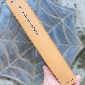 May include: A tan book spine with the title "Magick in Theory and Practice" by Crowley. The publisher is "Castle". The spine is set against a blurred, patterned background.