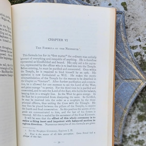 May include: An open book displaying the text "Chapter VI The Formula of the Neophyte." The pages are cream-coloured with black text. A decorative blue and gold corner piece is visible. The book is open to page 39.