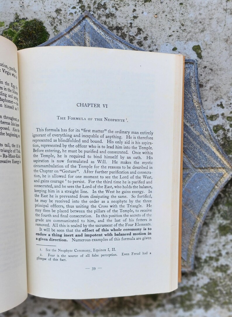 May include: An open book displaying the text "Chapter VI The Formula of the Neophyte." The pages are cream-coloured with black text. A decorative blue and gold corner piece is visible. The book is open to page 39.