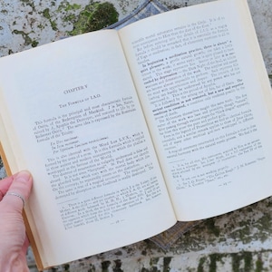 May include: An open book with aged, yellowed pages, displaying text from "CHAPTER V THE FORMULA OF I.A.O." The book is held open by a hand with a silver ring. The background is a textured, green and grey surface.
