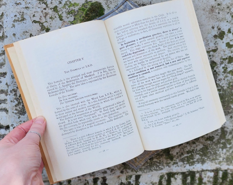 May include: An open book with aged, yellowed pages, displaying text from "CHAPTER V THE FORMULA OF I.A.O." The book is held open by a hand with a silver ring. The background is a textured, green and grey surface.