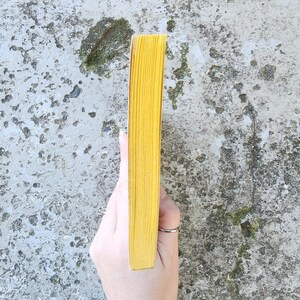 May include: A yellow book with a textured spine. The book's pages are a bright yellow colour. The book is held by a hand against a mottled grey background.