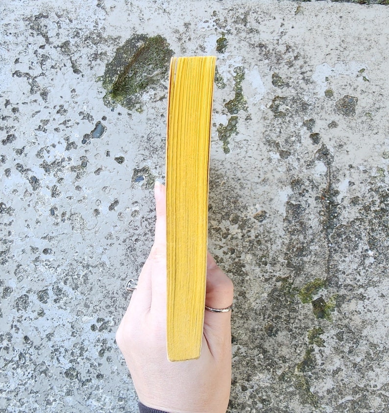 May include: A yellow book with a textured spine. The book's pages are a bright yellow colour. The book is held by a hand against a mottled grey background.