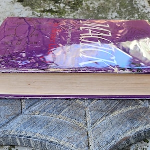 May include: A book titled "Magick" with a purple cover and white text, wrapped in clear plastic. The book rests on a grey stone surface. The pages are a light beige colour. The book is likely about magic.