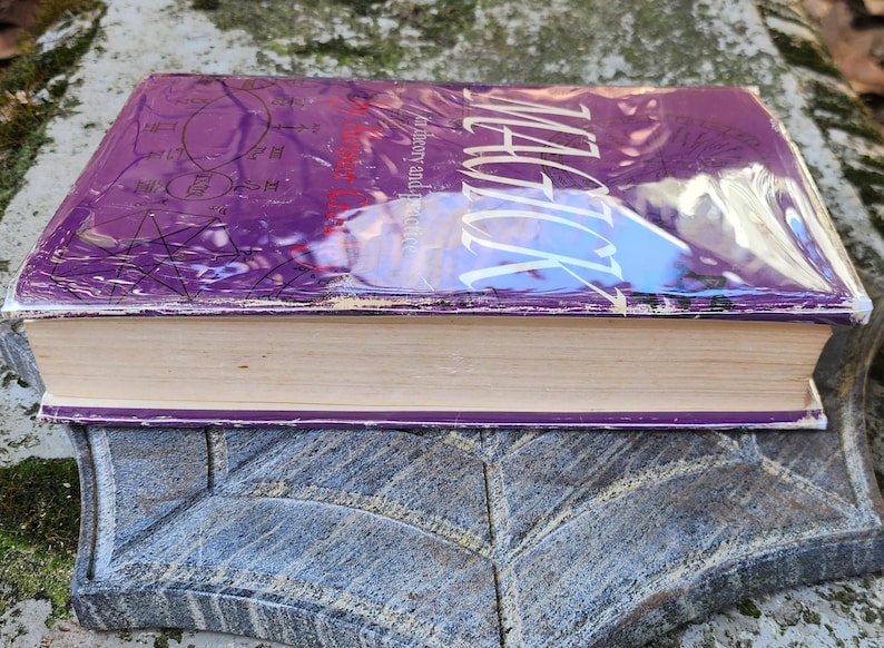 May include: A book titled "Magick" with a purple cover and white text, wrapped in clear plastic. The book rests on a grey stone surface. The pages are a light beige colour. The book is likely about magic.
