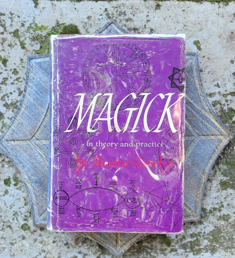 May include: A purple book with the title "MAGICK" in white, subtitled "in theory and practice", by Aleister Crowley. The cover has geometric illustrations and is wrapped in plastic. The book is placed on a decorative, star-shaped surface.