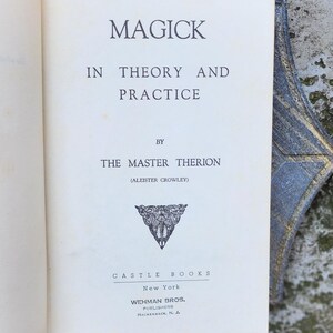 May include: An open book with the title "MAGICK IN THEORY AND PRACTICE" by The Master Therion (Aleister Crowley). The book has an off-white cover with black text and a decorative emblem. Published by Castle Books, New York.