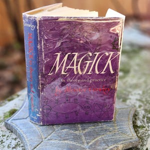 May include: A well-worn book titled "MAGICK in theory and practice" by Aleister Crowley. The purple cover features white lettering and is set on a grey, leaf-shaped stone. The book's spine is blue.