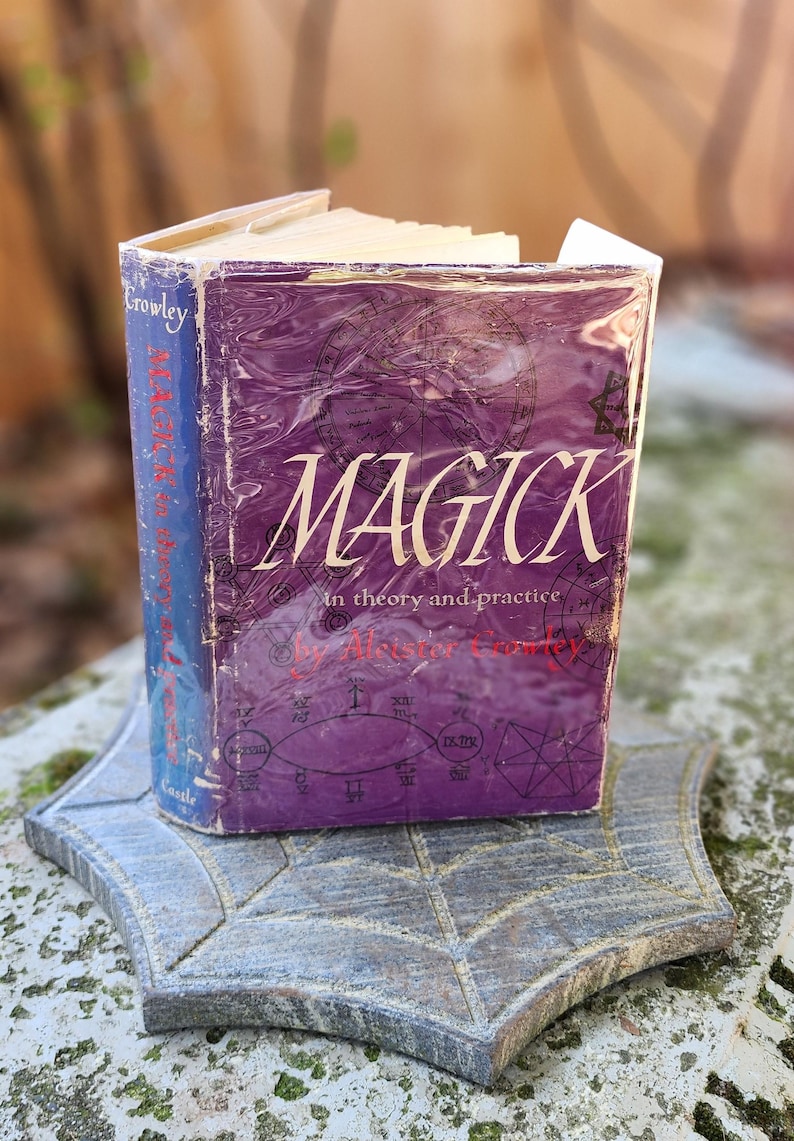 May include: A well-worn book titled "MAGICK in theory and practice" by Aleister Crowley. The purple cover features white lettering and is set on a grey, leaf-shaped stone. The book's spine is blue.