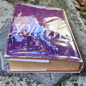 May include: A book with a purple cover and the word "MAGICK" in white lettering. The book is resting on a grey, textured surface. The book's pages are yellowed. The book appears to be old.