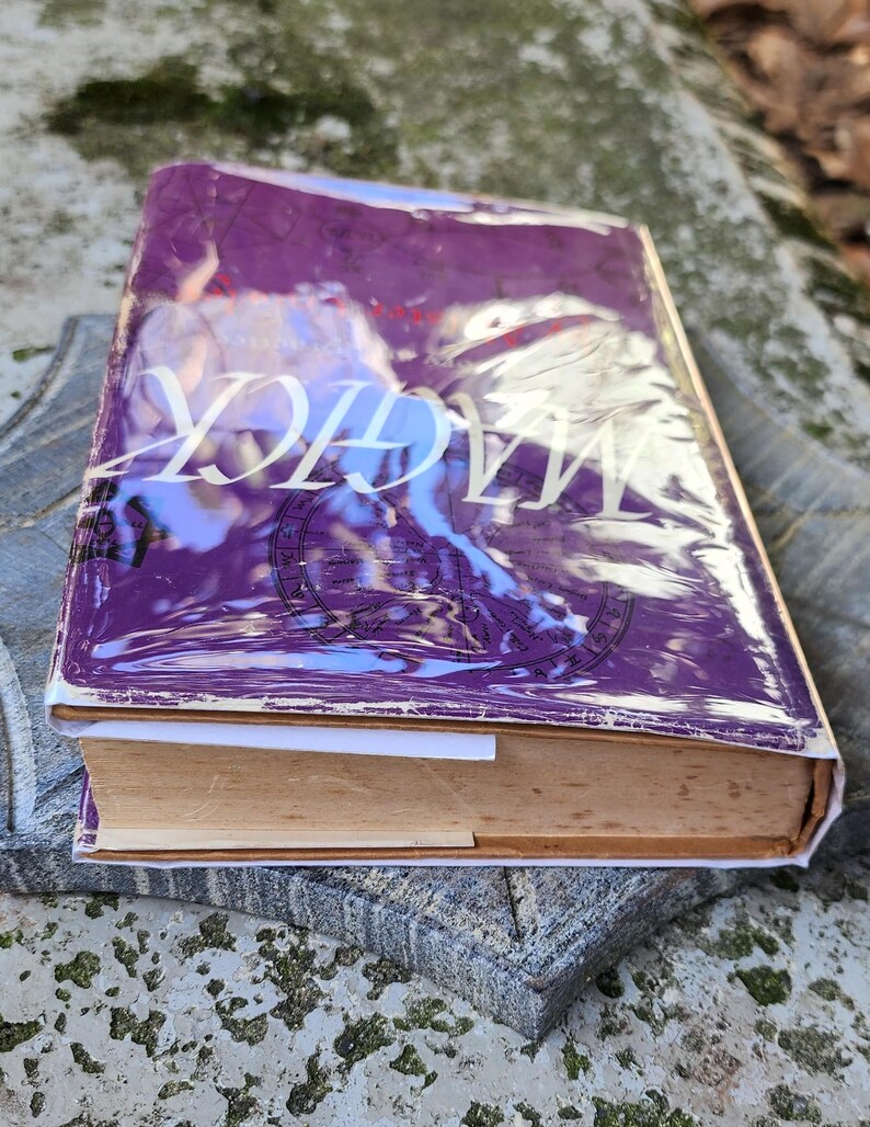 May include: A book with a purple cover and the word "MAGICK" in white lettering. The book is resting on a grey, textured surface. The book's pages are yellowed. The book appears to be old.