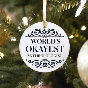 May include: White round Christmas ornament with black text and decorative flourishes. The text reads "World's Okayest Anthropologist".