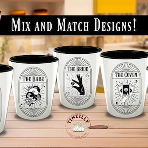 Op de afbeelding: Vijf shotglazen met zwarte randen en witte binnenkant. Elk glas heeft een ander ontwerp met zwart-wit illustraties. De ontwerpen omvatten "The Babe", "The Bride" en "The Coven".